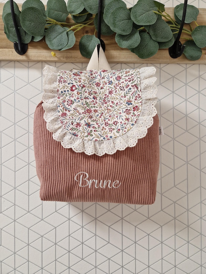 Puede incluir: Una mochila de pana color rosa empolvado con una solapa de estampado floral y ribete de encaje blanco. El nombre Brune est&aacute; bordado en cursiva blanca en la parte delantera. La mochila incluye un asa superior y una peque&ntilde;a etiqueta lateral.