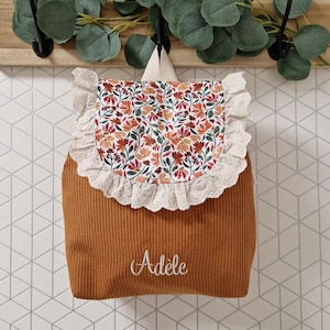 Puede incluir: Una mochila de pana color amarillo mostaza con una solapa de estampado floral y un borde de encaje blanco con ojales. El nombre Ad&egrave;le est&aacute; bordado en cursiva blanca en la parte frontal. La mochila tiene dos correas de hombro beige claro.