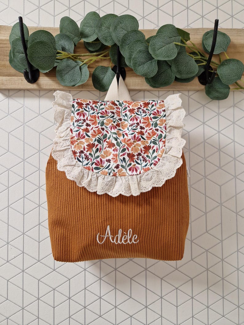 Puede incluir: Una mochila de pana color amarillo mostaza con una solapa de estampado floral y un borde de encaje blanco con ojales. El nombre Ad&egrave;le est&aacute; bordado en cursiva blanca en la parte frontal. La mochila tiene dos correas de hombro beige claro.