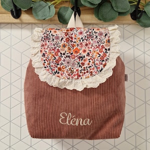 Puede incluir: Una mochila de pana color rosa empolvado con una solapa redondeada que presenta un estampado floral colorido y un borde de encaje color crema. El nombre El&eacute;na est&aacute; bordado en blanco en la parte frontal de la bolsa, que incluye un asa superior.