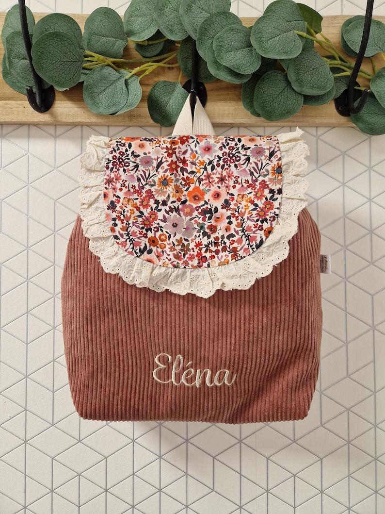 Puede incluir: Una mochila de pana color rosa empolvado con una solapa redondeada que presenta un estampado floral colorido y un borde de encaje color crema. El nombre El&eacute;na est&aacute; bordado en blanco en la parte frontal de la bolsa, que incluye un asa superior.