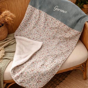 Peut inclure: Une couverture douce avec un motif floral composé de petites fleurs rouges, bleues et vertes sur fond blanc. La partie supérieure est en tissu côtelé bleu-gris uni avec le prénom Garance brodé en lettres cursives blanches.