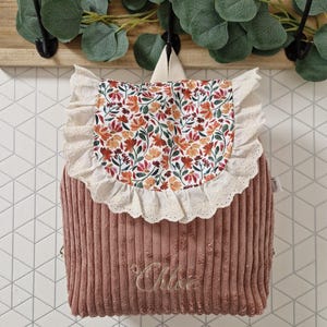 Puede incluir: Una mochila de pana color rosa empolvado con una solapa de estampado floral en tonos naranja, rojo y verde. La solapa est&aacute; bordeada con un volante de encaje color crema. El nombre Chloe est&aacute; bordado en letra cursiva en la parte frontal.