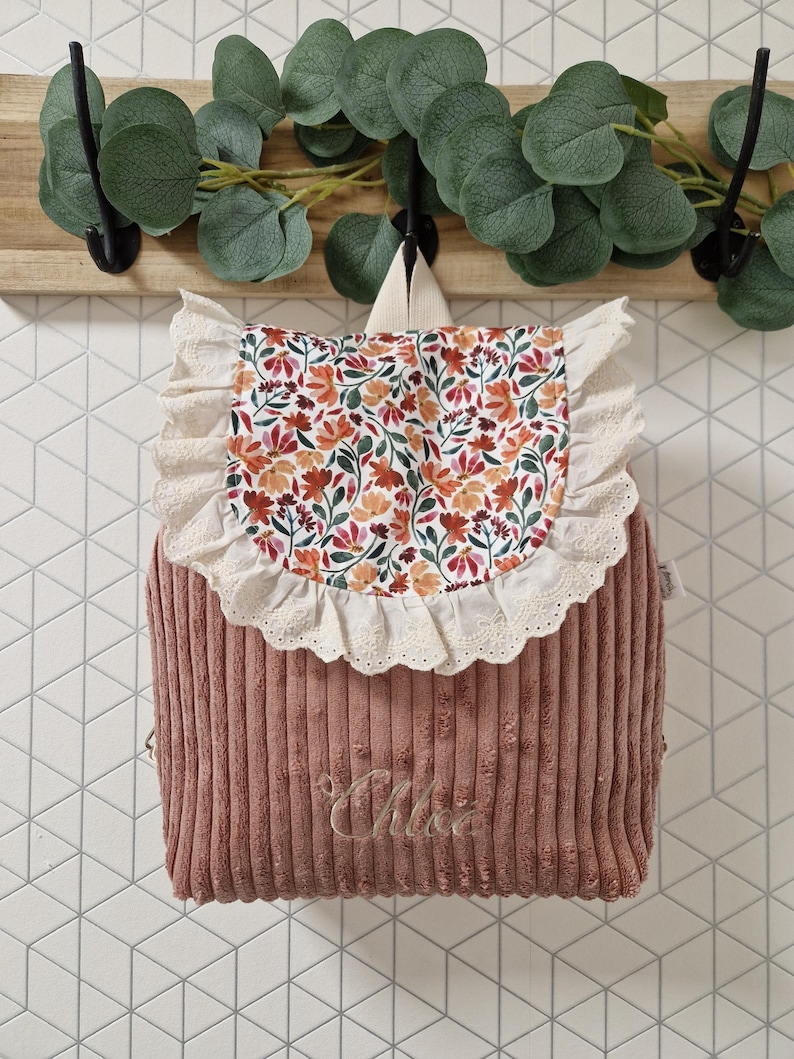 Puede incluir: Una mochila de pana color rosa empolvado con una solapa de estampado floral en tonos naranja, rojo y verde. La solapa est&aacute; bordeada con un volante de encaje color crema. El nombre Chloe est&aacute; bordado en letra cursiva en la parte frontal.