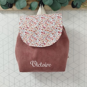 Peut inclure: Un sac à dos en velours côtelé rose poudré avec un rabat à motifs floraux. Le rabat présente un fond blanc avec un motif de fleurs et de feuilles rouges, oranges et grises. Le nom "Victoire" est brodé en blanc.