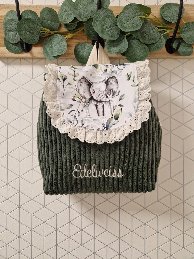 Puede incluir: Una mochila de pana verde oscuro con una solapa estampada con un elefante y follaje, adornada con encaje de ojales color crema. El nombre Edelweiss est&aacute; bordado en letra cursiva blanca en la parte frontal de la tela de pana.