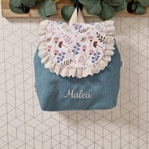 Puede incluir: Una mochila de pana azul con una solapa blanca que presenta un estampado floral en rosa, azul y marr&oacute;n. La solapa est&aacute; adornada con encaje de ojales color crema. El nombre Malea est&aacute; bordado en letra cursiva blanca en la parte frontal.