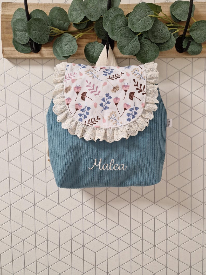 Puede incluir: Una mochila de pana azul con una solapa blanca que presenta un estampado floral en rosa, azul y marr&oacute;n. La solapa est&aacute; adornada con encaje de ojales color crema. El nombre Malea est&aacute; bordado en letra cursiva blanca en la parte frontal.