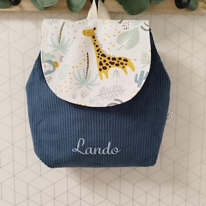 Puede incluir: Una mochila de pana azul marino con una solapa de fondo blanco y un estampado de jirafa y hojas de selva. El nombre Lando est&aacute; bordado en letra cursiva blanca en la parte frontal de la bolsa. Incluye un asa superior y correas para los hombros.