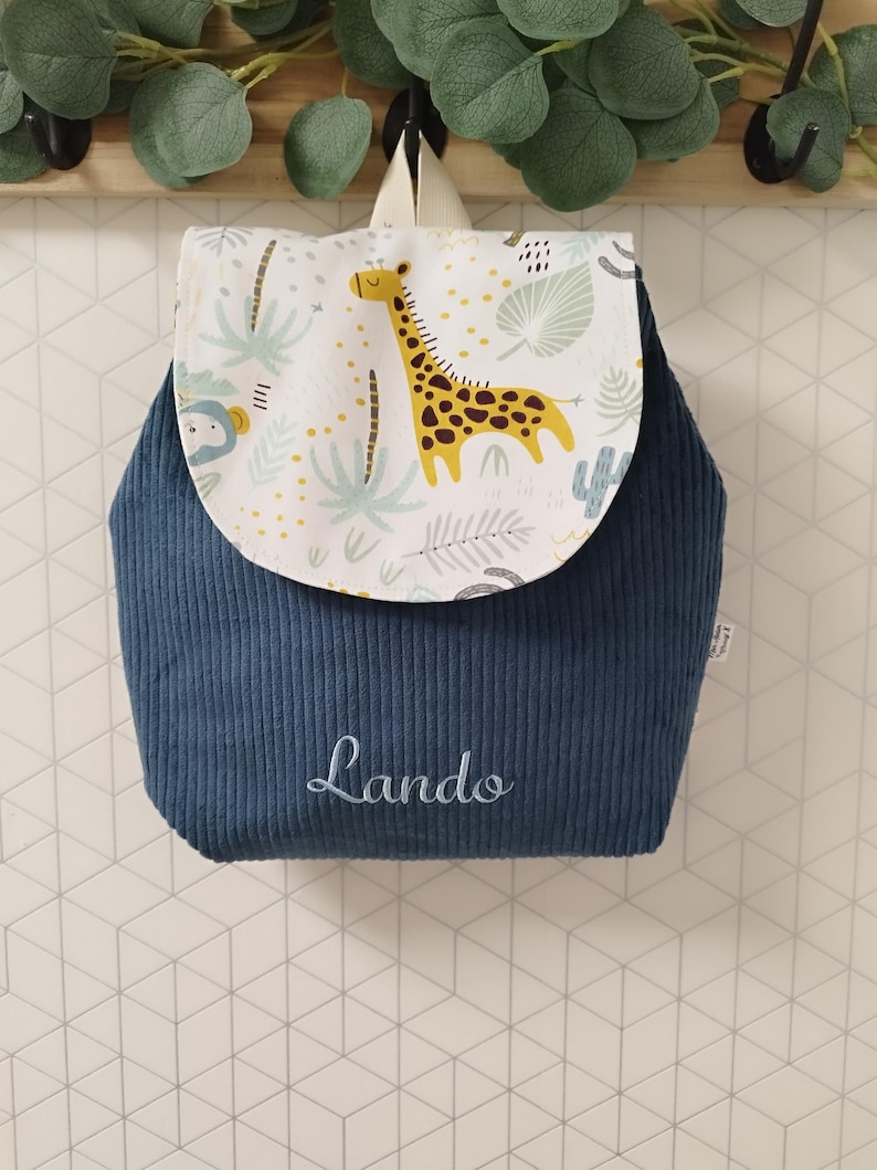 Puede incluir: Una mochila de pana azul marino con una solapa de fondo blanco y un estampado de jirafa y hojas de selva. El nombre Lando est&aacute; bordado en letra cursiva blanca en la parte frontal de la bolsa. Incluye un asa superior y correas para los hombros.