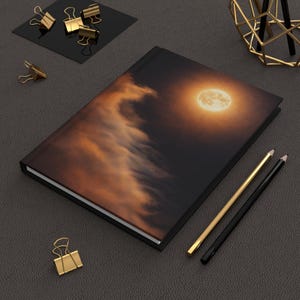 May include: A hardcover journal with a celestial design featuring a full moon and clouds in shades of orange and black. Gold and black pencils and gold binder clips are arranged around the journal.