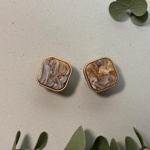 Könnte beinhalten: Ein Paar quadratische Ohrstecker mit einem marmorierten Muster in Beige-, Grau- und Cremetönen, eingefasst in goldfarbenem Metall. Die Ohrringe werden auf einer hellgrauen Oberfläche mit grünen Zierblättern präsentiert.