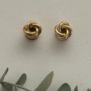 Könnte beinhalten: Ein Paar goldfarbene Ohrstecker mit einem polierten, ineinander verschlungenen Knotendesign. Jeder Ohrring besteht aus mehreren Metallschleifen, die eine kreisförmige, dreidimensionale Form bilden. Die Ohrringe haben eine glatte, reflektierende Oberfläche.