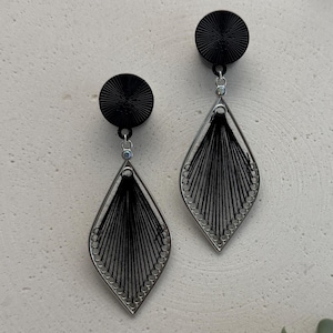 Könnte beinhalten: Ein Paar schwarz-silberner Ohrringe. Jeder Ohrring hat einen schwarzen, kreisförmigen Stecker, der mit einem silberfarbenen, rautenförmigen Rahmen verbunden ist, der mit schwarzen Fäden gefüllt ist. Die Ohrringe haben ein modernes, geometrisches Design.