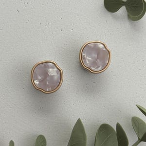 Könnte beinhalten: Zwei dekorative Schrankknöpfe. Jeder Knopf hat eine hellrosa, unregelmäßig geformte Mitte mit einem goldfarbenen Rand. Die Knöpfe sind vor einem hellgrauen Hintergrund mit grünen Blättern angeordnet.