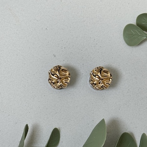 Könnte beinhalten: Ein Paar goldfarbene Ohrstecker mit einem strukturierten, floralen Design. Die Ohrringe sind rund und haben ein leicht erhabenes Muster. Der Hintergrund ist hellgrau mit grünen Blättern.