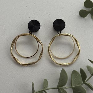 Könnte beinhalten: Ein Paar Ohrringe mit einem schwarzen, runden Stecker, der ein texturiertes Muster aus radialen Linien aufweist. Jeder Stecker ist mit zwei hängenden goldfarbenen Ringen verbunden: einem glatten, kleineren Innenring und einem größeren Außenring mit gewelltem Rand.