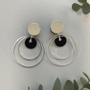 Könnte beinhalten: Ein Paar silberfarbene Creolen mit einer schwarzen Scheibe und einem kleinen, klaren Edelstein. Die Ohrringe haben ein kreisförmiges Design mit einer silbernen Scheibe oben. Moderner, minimalistischer Stil.