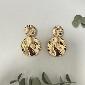 Könnte beinhalten: Ein Paar goldfarbene Ohrringe mit einem Doppel-Tropfen-Design. Jeder Ohrring besteht aus einem kleineren runden oberen Teil und einem größeren runden unteren Teil, beide mit einer gehämmerten, reflektierenden und unebenen Metalloberfläche.