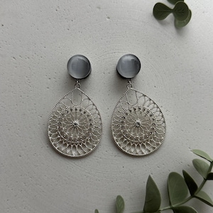 May include: A pair of silver-toned, filigree teardrop earrings with gray cabochon studs. The earrings feature intricate, openwork designs with a central, circular pattern and small, sparkling accents. The earrings are displayed on a light gray surface.