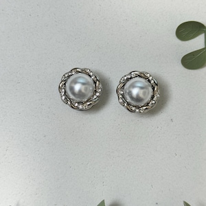May include: Two pearl stud earrings. Each earring has a large, round, white pearl encircled by a silver-coloured, twisted rope design and a ring of small, clear crystals. The earrings are set against a light grey background.