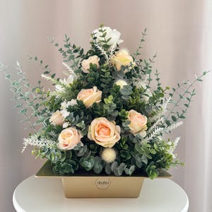 Könnte beinhalten: Blumenarrangement mit pfirsichfarbenen Rosen, weißen Blüten und verschiedenen grünen Blättern, präsentiert in einem goldfarbenen rechteckigen Behälter. Die Anordnung steht auf einer weißen Oberfläche. Der Behälter trägt das Wort "GINARIA".