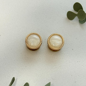 May include: Two round, gold-toned earrings with a cream-colored, marbled center. The earrings have a textured, beaded border. The background is a light, neutral color with green sprigs in the corners.