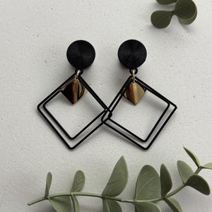 Op de afbeelding: Zwarte en gouden geometrische oorbellen. Elke oorbel heeft een zwarte cirkelvormige stud, een goudkleurige diamantvormige bedel en een zwart dubbel vierkant frame. Gepresenteerd op een wit oppervlak met groene bladaccenten.