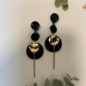 Könnte beinhalten: Ein Paar schwarze und goldfarbene Hängeohrringe. Jeder Ohrring hat ein strukturiertes schwarzes Oberteil, gefolgt von einer schwarzen Scheibe, einer goldenen Scheibe und einem goldenen Stab. Heller Hintergrund.