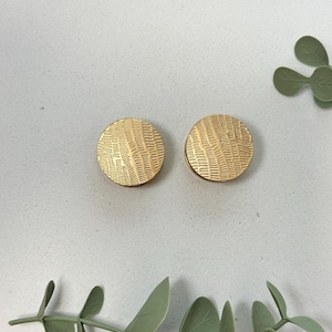 Könnte beinhalten: Zwei goldfarbene, runde Ohrstecker mit einem strukturierten, linearen Muster. Die Ohrringe werden auf einer weißen Oberfläche mit grünen Blättern im Hintergrund präsentiert. Die Ohrringe sind ein Schmuckstück.