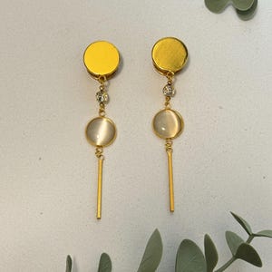 Könnte beinhalten: Ein Paar goldfarbene Ohrringe mit kreisförmigem Oberteil und hängendem Design. Jeder Ohrring hat einen klaren Kristallakzent und einen runden, cremefarbenen Stein. Unterhalb des Steins hängt ein dünner, goldener Stab. Die Ohrringe werden auf einer hellgrauen Oberfläche präsentiert.