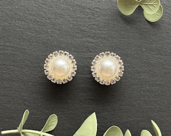 May include: A pair of circular stud earrings featuring a central white faux pearl surrounded by a halo of small, clear, sparkling crystals. The earrings have a metallic setting and are displayed alongside decorative light green plastic leaves.