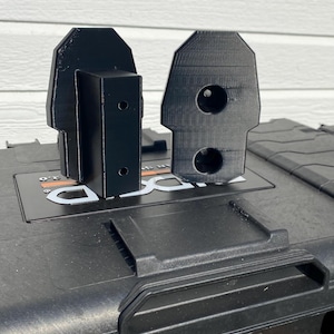 Puede incluir: Dos soportes negros impresos en 3D con un diseño geométrico se muestran encima de un contenedor de almacenamiento negro. Los soportes tienen agujeros y están diseñados para sujetarse al contenedor. El contenedor tiene un asa naranja.