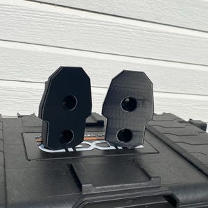 May include: Two black, 3D-printed brackets with a rectangular shape and rounded top. Each bracket has two circular holes. The brackets are displayed on a black case with a white background.