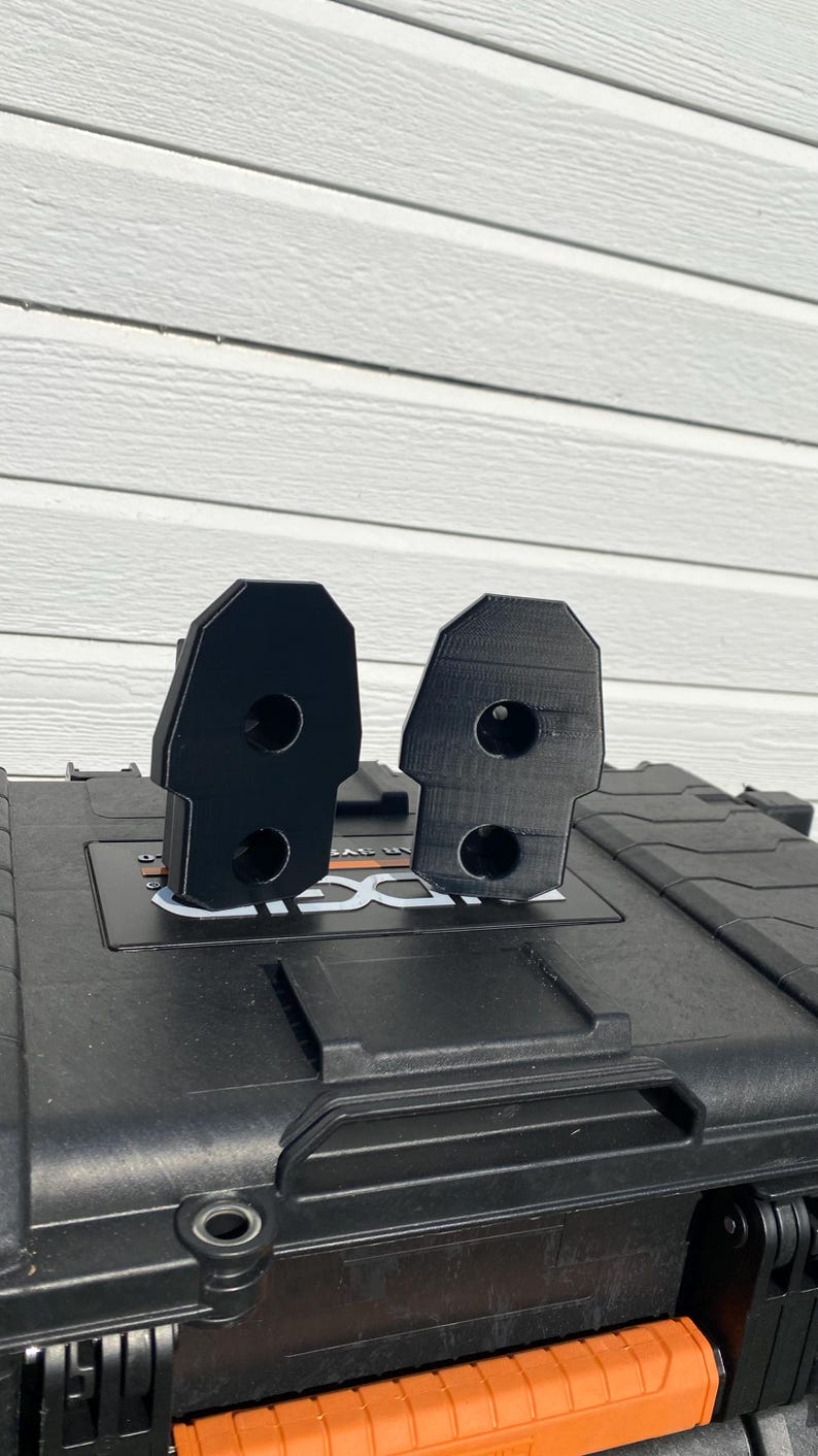 May include: Two black, 3D-printed brackets with a rectangular shape and rounded top. Each bracket has two circular holes. The brackets are displayed on a black case with a white background.