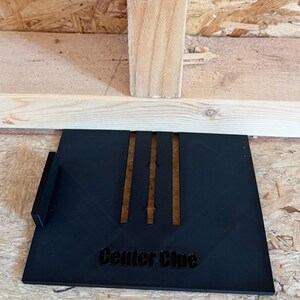 Puede incluir: Herramienta de plástico negro con el texto "Center Clue" y tres ranuras paralelas. La herramienta está diseñada para ayudar a centrar. El fondo es una estructura de madera.