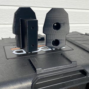 May include: Two black, 3D-printed brackets are displayed on top of a black storage container. The brackets have a unique, geometric design with holes and a rectangular shape. The container has an orange handle.