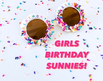 May include: White plastic sunglasses with brown lenses decorated with colorful sprinkles and the words "GIRL" and "BIRTHDAY".