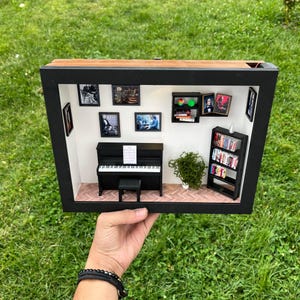 Può includere: Diorama di una stanza in miniatura con un pianoforte nero, spartiti e una piccola panca nera. Le pareti espongono opere d'arte incorniciate e una piccola libreria è piena di libri in miniatura. Il diorama è in una cornice nera con un piano in legno.