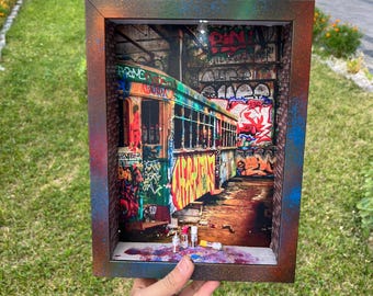 May include: A framed artwork showing a graffiti-covered tram in a tunnel. The artwork features vibrant colours and detailed graffiti art. The frame is a dark brown with a multi-coloured border. Small paint bottles are in front of the artwork.