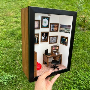 Puede incluir: Un diorama en miniatura de una escena de oficina en casa, enmarcado en negro y madera. El interior presenta un escritorio, una silla, un ordenador portátil y obras de arte enmarcadas. Un puf rojo está en la esquina. El diorama se sostiene contra un fondo de hierba verde.