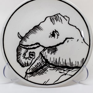 Può includere: Un piatto in ceramica bianca con un disegno in bianco e nero di una testa di elefante. La proboscide dell'elefante è arrotolata e il suo occhio è visibile.