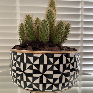 Op de afbeelding: Een zwarte en witte geometrische gepatenteerde keramische plantenpot met drie zwarte poten. De pot is gevuld met een cactusplant.