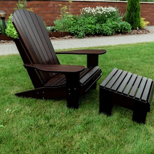 Op de afbeelding: Een donkerbruine houten Adirondack-stoel met een bijpassende voetenbank. De stoel en voetenbank staan op een groen gazon.