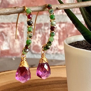 May include: A pair of gold hoop earrings with a teardrop-shaped pink gemstone dangling from each hoop. The hoops are made of small green and red beads.
