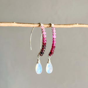 May include: A pair of silver hoop earrings featuring a gradient of pink and red gemstones. Each earring has a teardrop-shaped moonstone pendant.