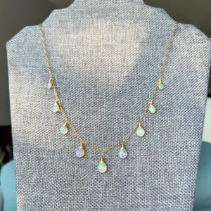 May include: A delicate gold necklace featuring multiple opal pendants. The opals have a pearlescent sheen, and the necklace is displayed on a gray fabric background. The chain is thin and elegant, with the pendants evenly spaced.