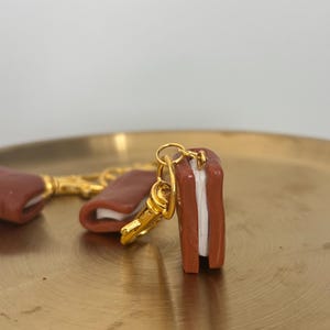 Op de afbeelding: Een miniatuur sleutelhanger in de vorm van een bruin en wit sandwich met een goudkleurige sluiting. De sandwich heeft een rechthoekige vorm met een witte vulling tussen twee bruine broodjes. De sleutelhanger ligt op een goudkleurig oppervlak.