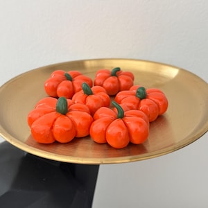 Op de afbeelding: Zes kleine, oranje pompoenen met groene stelen liggen op een goudkleurig dienblad. De pompoenen zijn fel oranje en lijken glad. De ronde schaal heeft een glanzende afwerking, wat een feestelijke herfstpresentatie creëert.