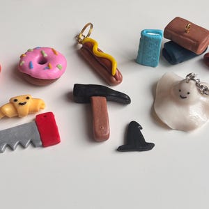 Op de afbeelding: Een verzameling miniatuur, handgemaakte bedels in verschillende vormen en kleuren. Inclusief een pompoen, donut, croissant, zaag, hotdog, hamer, heksenhoed, spook en kleine boeken. Sommige bedels zijn bevestigd aan sleutelhangers.